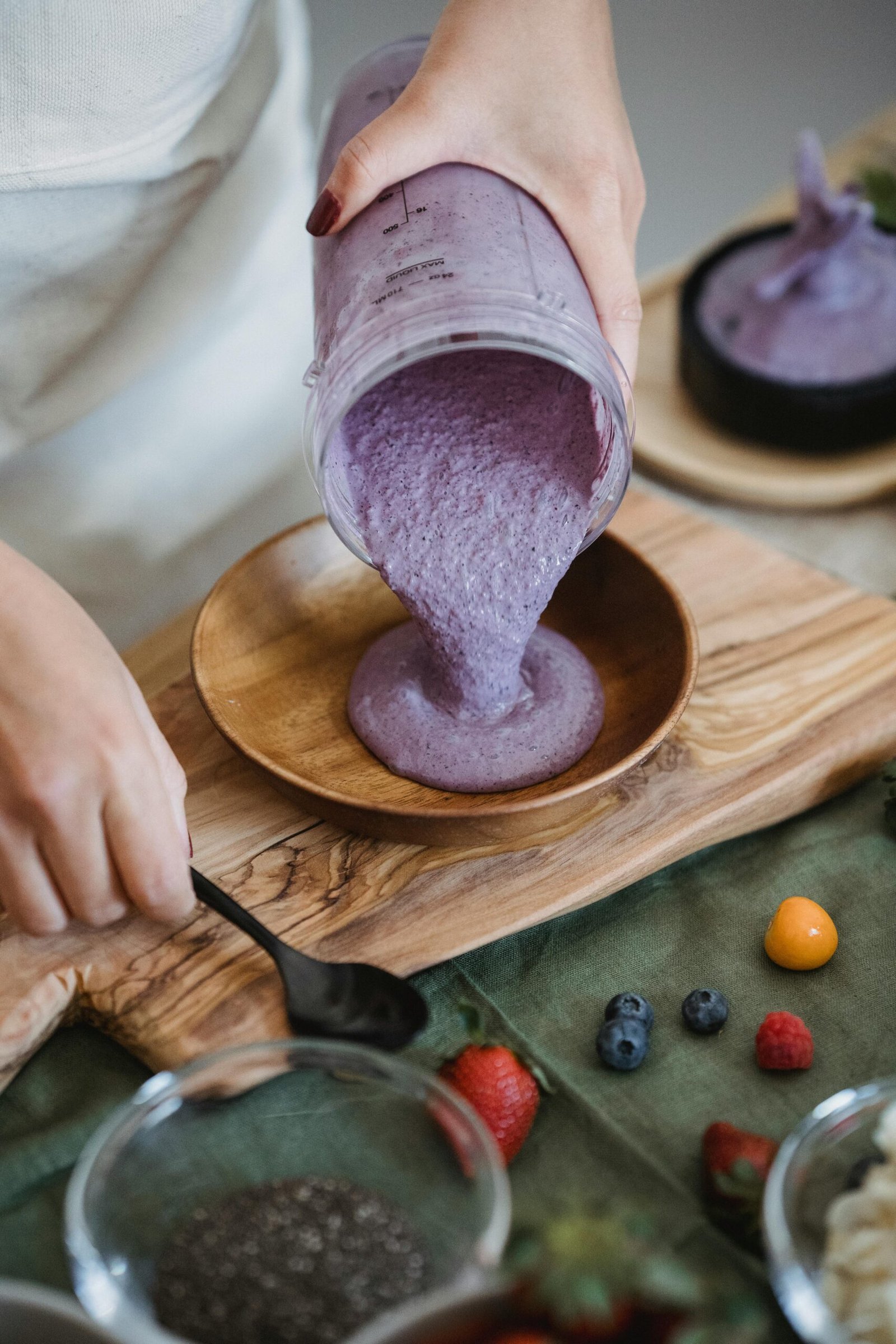 Close-up of a berry smoothie being poured into a wooden bowl, surrounded by fresh ingredients.
